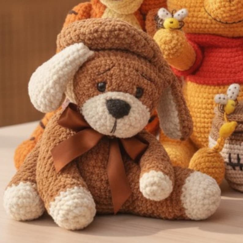 May include: A brown and cream crocheted dog plush toy featuring a dark brown satin ribbon tied around its neck, floppy ears, and a dark nose. The toy is crafted with a textured yarn finish and is positioned in front of other crocheted figures.