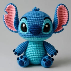 May include: A crochet amigurumi doll of a blue creature with large ears, pink inner ear details, and a light blue belly. The doll features a dark blue nose, large black eyes with white highlights, and small dark blue claws on its hands and feet.