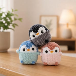 May include: Three small, round, crocheted plush toys shaped like penguins. One is dark gray and white, one is light blue and white, and one is light pink and white. Each penguin features black plastic eyes, a small orange beak, and tiny wings.
