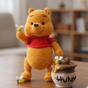 May include: A yellow crocheted bear figure wearing a red shirt, holding a small bee on one hand. Beside the bear is a tan and brown crocheted honey pot with a lid, featuring the word HUNNY embroidered in black thread and yellow yarn drips.