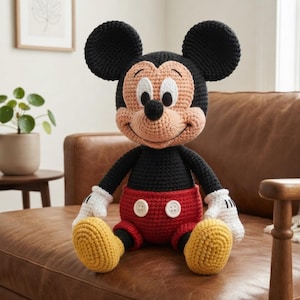 Op de afbeelding: Een handgehaakte pop van Mickey Mouse met een zwart hoofd en romp, grote ronde oren, een perzikkleurig gezicht, witte handschoenen, een rode broek met twee witte knopen en gele schoenen. Het figuurtje heeft een opgestikte glimlach.