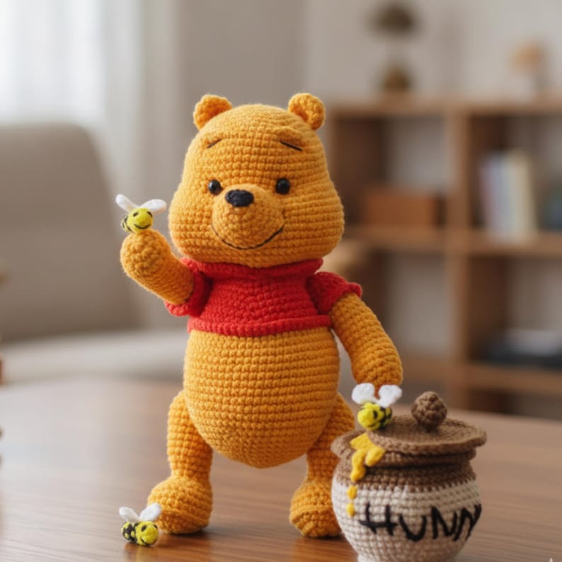 May include: A yellow crocheted bear toy wearing a red shirt, holding a small crocheted bee in one raised hand. Beside the bear is a brown crocheted honey pot with a lid, featuring yellow yarn dripping down the side and the word HUNNY stitched on it.