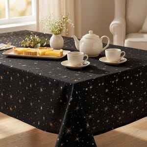 May include: A black tablecloth featuring a gold star pattern, set with a white ceramic teapot, two matching teacups on saucers, and a rectangular white platter holding four slices of yellow tart, alongside a small white vase with white flowers.