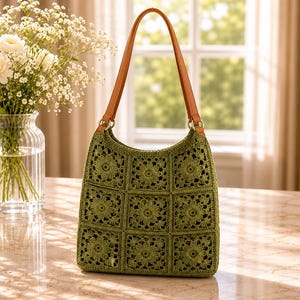 May include: A sage green crochet handbag constructed from nine square floral motifs arranged in a grid. The bag features two tan leather-like shoulder straps attached with gold-toned metal rings, providing a structured shape for the soft textile body.