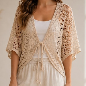 May include: A beige crochet cardigan featuring an open-knit pattern, short wide sleeves, and a front tie closure. It is worn over a plain white tank top paired with beige wide-leg trousers that have an elasticated drawstring waistband.
