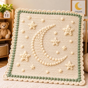 May include: A square crochet blanket in cream yarn featuring a central crescent moon and star pattern created with raised bobble stitches. The blanket is bordered by a sage green bobble trim and a scalloped cream edge, providing a textured finish.