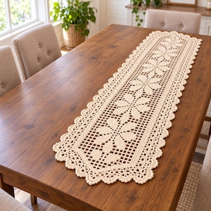Puede incluir: Un camino de mesa rectangular largo de ganchillo en color crema, con un patrón floral repetitivo, detalles de rejilla calada y un borde festoneado. El camino está hecho de hilo y muestra una serie de motivos florales interconectados.