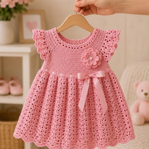 May include: A light pink crochet dress featuring flutter sleeves, a round neckline, and a textured stitch pattern. The bodice is adorned with a crocheted flower with a central pearl bead, and a satin ribbon bow is tied at the waistline.