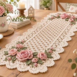 May include: An off-white rectangular crochet table runner featuring an intricate openwork lattice pattern and scalloped edges. The runner is decorated with three-dimensional crocheted pink roses and sage green leaves at both ends.