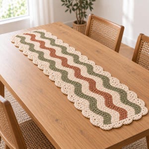 Può includere: Un runner da tavola rettangolare all'uncinetto caratterizzato da un motivo a onde con strisce alternate color crema, verde oliva e terracotta. I bordi del runner sono rifiniti con una bordura smerlata in filato color crema.