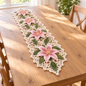 Puede incluir: Un camino de mesa de ganchillo color crema con un patrón repetitivo de cuatro grandes flores de lirio rosas tridimensionales con centros amarillos, rodeadas de hojas de ganchillo verde oscuro y pequeños capullos rosas en un borde festoneado.