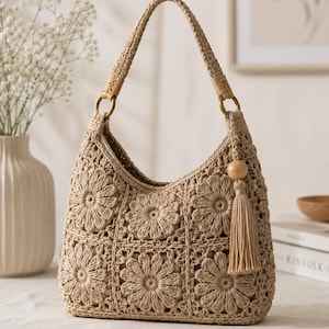 May include: A beige crochet handbag featuring a repeating floral motif pattern. The bag has a single braided handle attached with gold-toned metal rings and a decorative tassel accent with a wooden bead hanging from the right side of the opening.