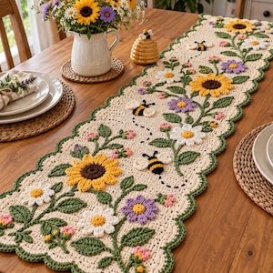 May include: A cream-colored crochet table runner featuring a scalloped green border and a floral pattern with yellow sunflowers, white daisies, purple flowers, and small pink buds. Three-dimensional bees are stitched along the runner.
