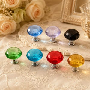 Puede incluir: Un conjunto de siete pomos para armarios con forma de diamante y facetas, hechos de vidrio translúcido de colores, incluyendo azul, morado, negro, verde, turquesa, rojo y amarillo, cada uno fijado a una base circular de metal plateado pulido.