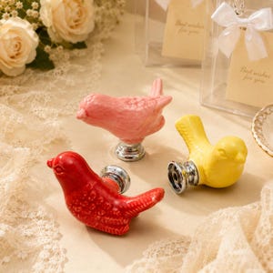 May include: Three ceramic bird-shaped drawer knobs in red, pink, and yellow. Each bird features a glossy finish and is attached to a circular silver-toned metal base. The knobs are designed for furniture hardware, showcasing detailed molded feathers.