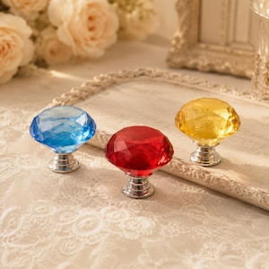 May include: Three faceted cabinet knobs with a diamond-like shape, each featuring a polished silver-tone metal base. The set includes one blue, one red, and one yellow translucent knob, all designed with a multi-faceted surface for light reflection.