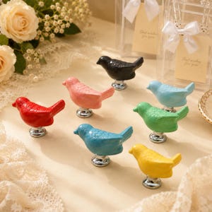 May include: A collection of seven bird-shaped cabinet knobs in various colors including red, pink, black, light blue, green, teal, and yellow. Each ceramic bird figure is mounted on a circular silver-toned metal base for attachment to furniture.