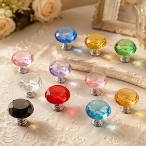 Puede incluir: Una colección de once pomos para armarios con forma de diamante y facetas en varios colores translúcidos, incluyendo azul, amarillo, verde, rosa, morado, rojo, transparente, negro y azul claro, con una base de metal plateado.