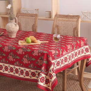 Puede incluir: Un mantel rectangular con una base de color rojo intenso y un patrón repetitivo de motivos florales en rosa apagado y follaje verde. Los bordes están rematados con una ancha cenefa de color crema decorada con flores rojo oscuro.