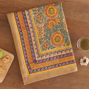 May include: A rectangular fabric tablecloth featuring a tan background with a vibrant sunflower print in yellow, orange, and green. The border displays a decorative pattern of yellow, blue, and red geometric shapes and floral motifs.