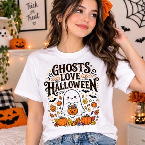 May include: A white short-sleeve t-shirt featuring a graphic print with the text GHOSTS LOVE HALLOWEEN in black and orange. The design includes a central white ghost holding a jack-o'-lantern, surrounded by orange pumpkins, autumn leaves, candy corn, and small black bats.