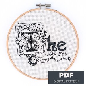 May include: Cross-stitch embroidery hoop featuring the word "The" in black thread. The letter "T" is ornate, within a decorative frame. The fabric is white, and the wooden hoop is visible. A PDF digital pattern is indicated.