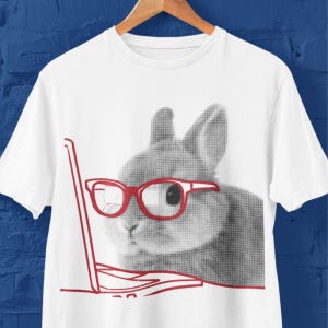 May include: A white short-sleeved t-shirt featuring a graphic print of a grey rabbit wearing bright red glasses. The rabbit is positioned behind a red outline of an open laptop computer. The design uses a dot-matrix style pattern for the rabbit.
