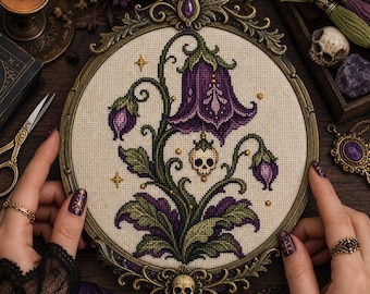 Può includere: Un ricamo a punto croce in una cornice ovale in bronzo antico raffigurante un fiore viola a forma di campana con foglie verdi e un piccolo teschio appeso al centro. Il disegno include piccole stelle dorate su un tessuto color crema.