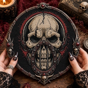 May include: A circular cross-stitch embroidery featuring a detailed beige skull with a cracked forehead and a dark metal-like jaw guard, set against a black background with red accents, framed in an ornate, dark metallic hoop with small skulls.