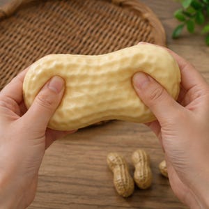 May include: A light beige, textured, peanut-shaped stress relief toy being squeezed. The surface of the toy features a bumpy, shell-like pattern that mimics the appearance of a real peanut. Two smaller, realistic peanut shells are visible below.