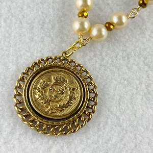 May include: Gold necklace with a large, round pendant featuring a gold crest with a crown and leaves. The necklace is made with white pearls and gold beads.