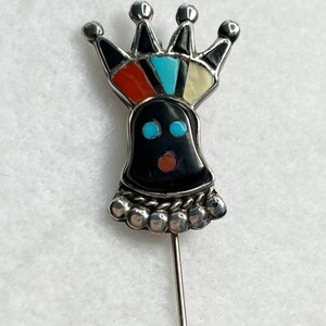 May include: A silver pin with a black, red, blue, and white stone design. The pin features a crown and a face with blue eyes and a red mouth.