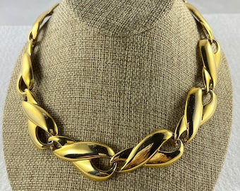 May include: A gold-toned statement necklace with large, twisted links. The necklace is displayed on a fabric-covered mannequin.
