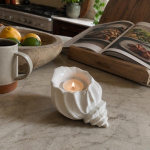 May include: A white ceramic candle holder shaped like a conch shell, featuring a glossy finish and a central circular opening that contains a small, lit tea light candle with a visible flame, resting on a smooth, light-colored stone surface.