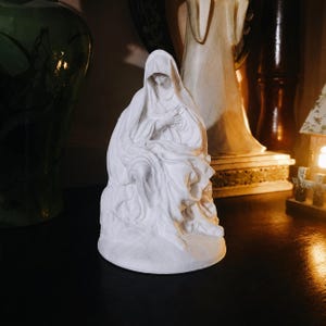 May include: A matte white sculpture of a seated figure draped in a hooded cloak. The figure is depicted in a contemplative posture with hands held near the chest, featuring intricate folds and deep shadows within the fabric of the garment.