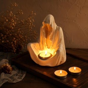 May include: A white, hooded, seated figure sculpture crafted from a matte material, designed to hold a small tea light candle in its lap. The figure features draped robes and a hollowed-out interior that glows with the warm light of the candle.