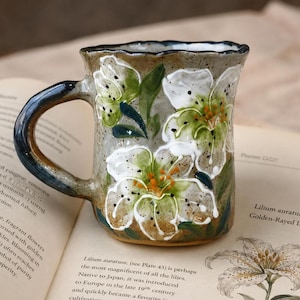 May include: A speckled ceramic mug with a wavy rim and a dark handle, featuring hand-painted white and green lily flowers with orange centers. The mug has a textured, earthy finish and is decorated with detailed floral motifs across its surface.