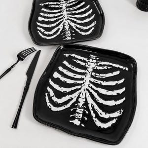 May include: Two square black plates featuring a white raised skeleton rib cage design. A black fork and a black knife are positioned to the left of the plates. The plates have rounded corners and a glossy finish, displaying a detailed human torso bone structure.