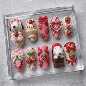 May include: A set of ten press-on nails featuring 3D designs including Snoopy figures, strawberries, cherries, red bows, chocolate bars, and heart-shaped frames. The color palette consists of soft pink, cream, red, and brown with intricate details.