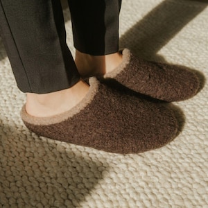 Puede incluir: Un par de zapatillas de lana afieltrada de color marrón oscuro con punta redondeada y forro interior de color beige claro. Las zapatillas presentan una superficie exterior texturizada y difusa con un diseño sin cordones, junto a un pantalón negro.