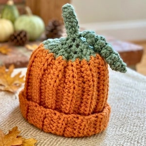 Peut inclure: Un bonnet crocheté de couleur orange citrouille vif, présentant une texture côtelée et un bord replié. Le sommet du bonnet est orné d'une tige crochetée vert sauge et d'un détail de vigne enroulée, imitant l'apparence d'une citrouille.