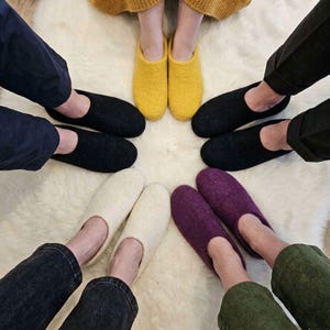 May include: A collection of felted wool slippers in various colours including mustard yellow, black, off-white, and deep purple. The slippers are arranged in a circular formation, each featuring a rounded toe, a low-cut opening, and a simple slip-on design.