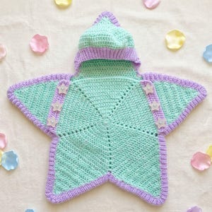 May include: A star-shaped crochet blanket with a hood, featuring a mint green center and a lavender border. The garment includes three small star-shaped buttons on each side, providing a decorative closure for this soft, textured textile piece.