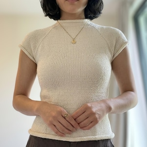 May include: A cream-coloured, short-sleeved knit top featuring a high mock neckline and a rolled hem finish. The garment is constructed with a fine-gauge knit texture and raglan-style sleeves, providing a fitted silhouette that sits at the waistline.