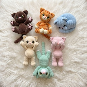 May include: Six crocheted cat plushies in various colors: a dark brown cat, an orange tabby, a curled-up light blue cat, a cream-colored cat, a mint green cat with an upright tail, and a pink cat with its paws covering its eyes.