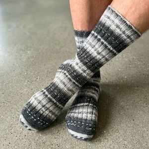 Puede incluir: Un par de calcetines de punto a media pantorrilla con un patrón jaspeado de hilos negros, gris carbón y blancos. Los calcetines tienen una textura acanalada en toda la pierna y el pie, con una puntera gris oscuro sólida y un aspecto moteado.