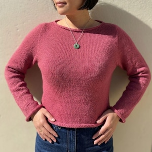 May include: A long-sleeved, crew-neck jumper in a solid dusty rose colour with a rolled hem at the neckline, cuffs, and bottom edge. The jumper is paired with dark blue denim jeans and a silver chain necklace featuring a round, light green pendant.