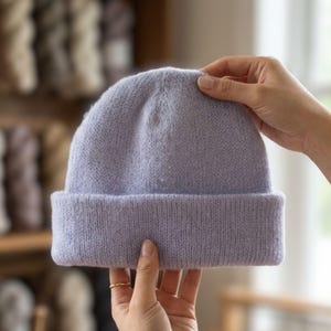 May include: A light lavender beanie made of a soft, fuzzy knit material. The hat features a wide, folded cuff at the bottom and a rounded crown, showcasing a consistent, uniform texture throughout the entire piece in a solid pastel purple color.