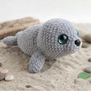 Peut inclure: Une peluche phoque en crochet gris avec une texture en fil. Le phoque présente de grands yeux ronds, brillants et verts avec des pupilles noires, ainsi qu'un petit nez noir lisse. Le corps est arrondi avec de petites nageoires et une queue.