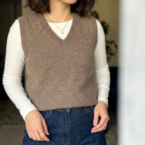 May include: A brown knit sweater vest with a V-neckline, worn over a long-sleeved white shirt and paired with dark blue denim jeans. The vest features a ribbed texture at the neckline and armholes, complemented by a simple gold-toned necklace.